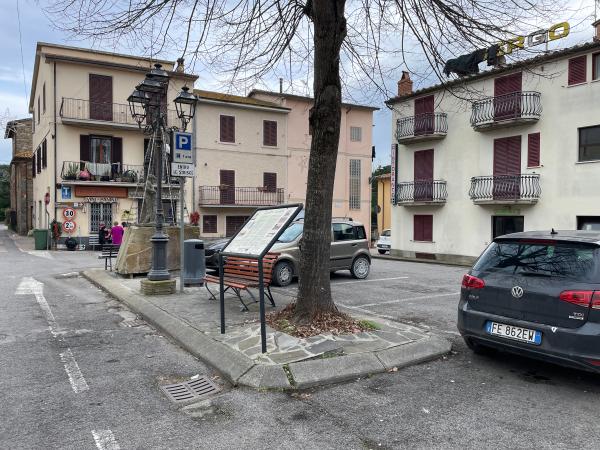 Place du Borghetto avec un arbre dénudé, un banc, un panneau d'information, un petit magasin, une fontaine et des balcons